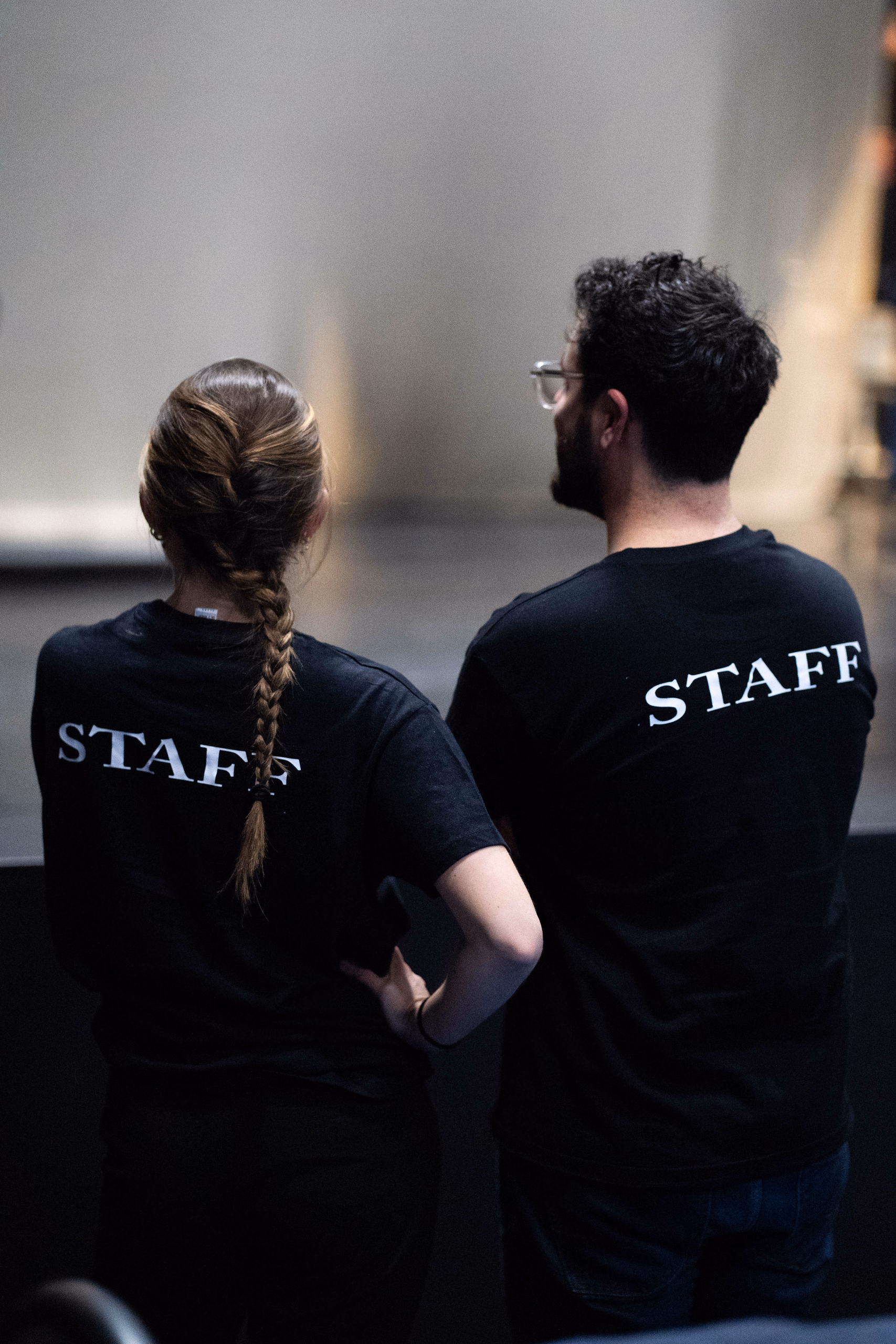 Deux membres de l'équipe, de dos, en t-shirt noir marqué « STAFF », regardent la scène