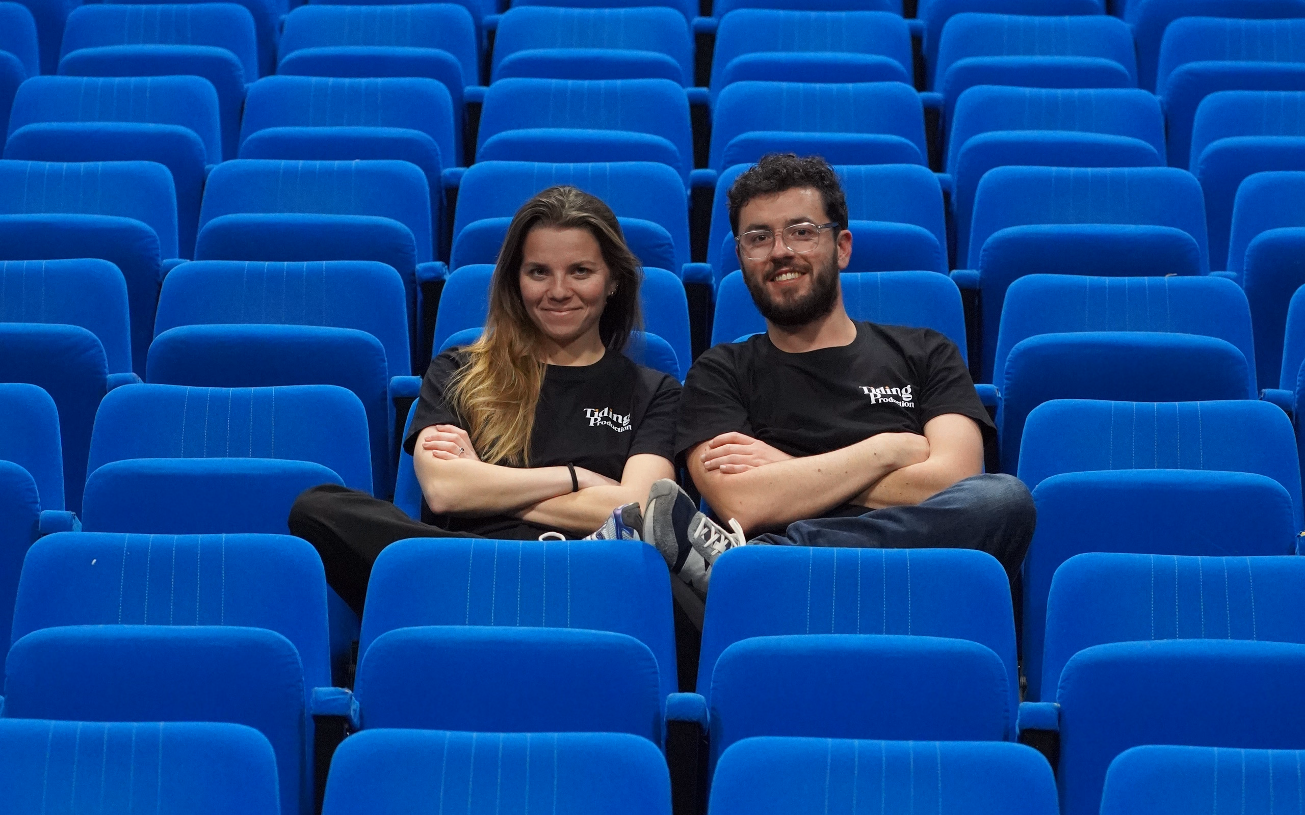 Deux membres de l'équipe Tiding Production sourient, bras croisés, au milieu de fauteuils de théâtre bleus vides