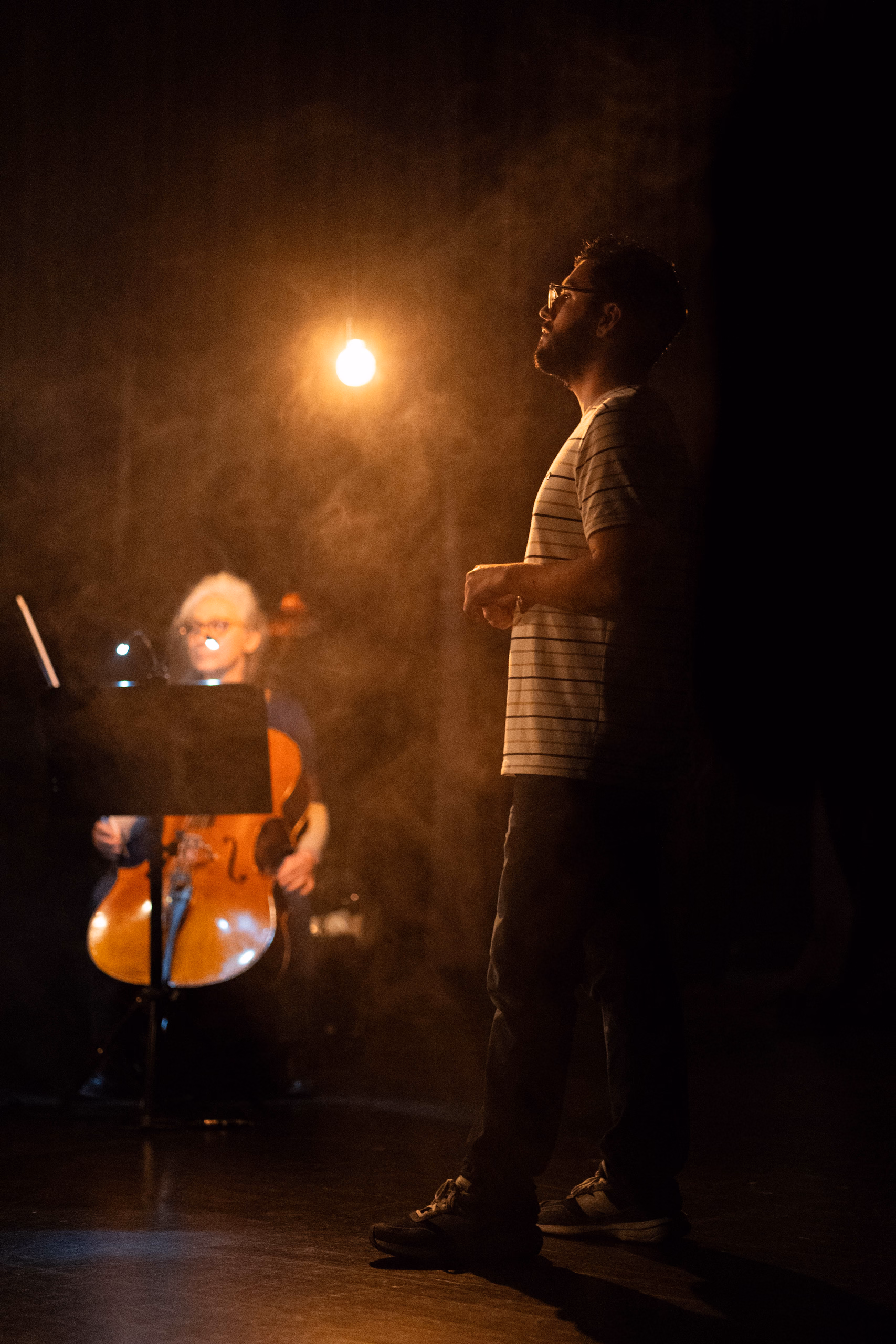 Dans la fumée et la lueur d'une ampoule, une personne en t-shirt rayé debout sur scène, violoncelle et pupitre en arrière-plan