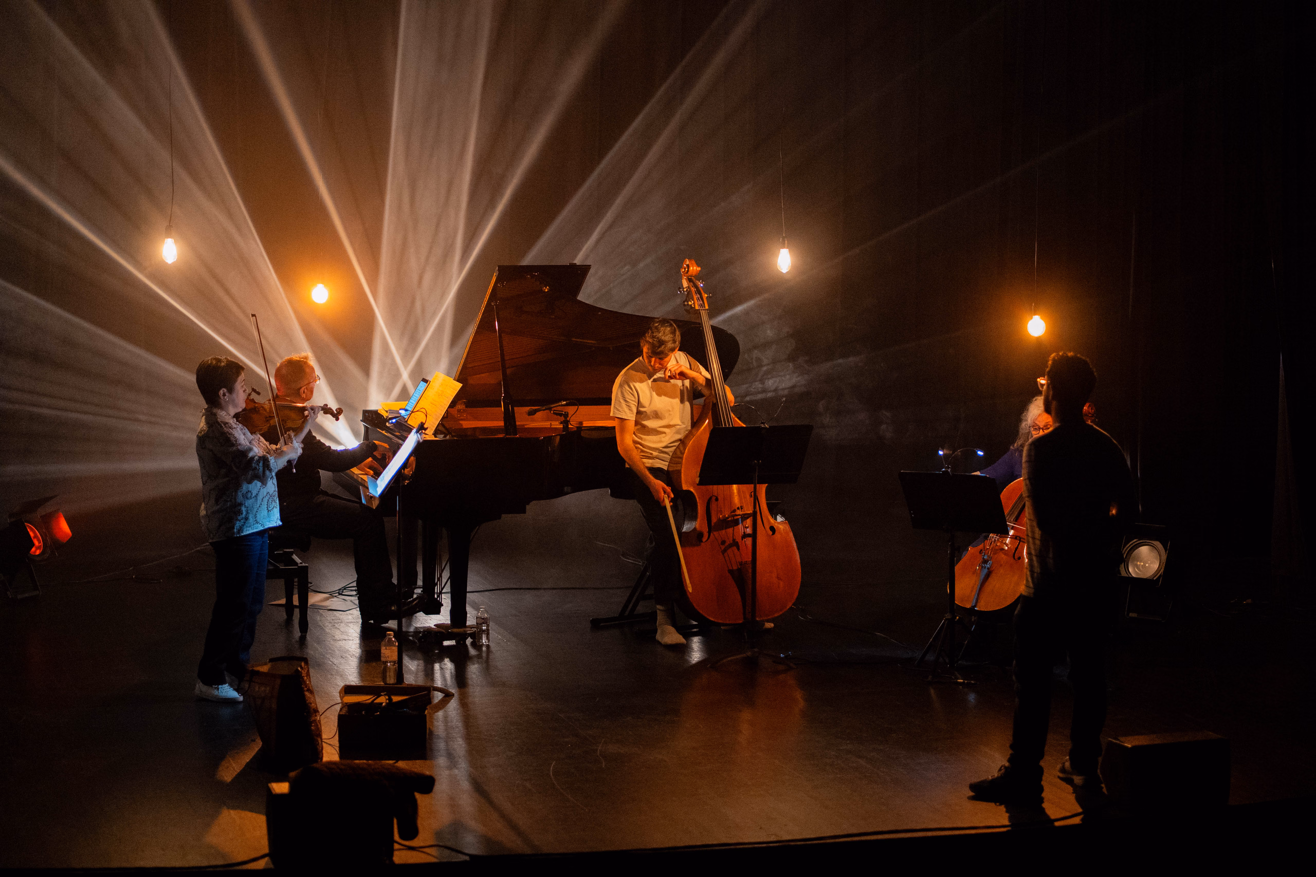 Musiciens en répétition sur scène (violon, piano à queue, contrebasse, violoncelle) sous des faisceaux de lumière et des ampoules suspendues, une personne debout de dos au premier plan