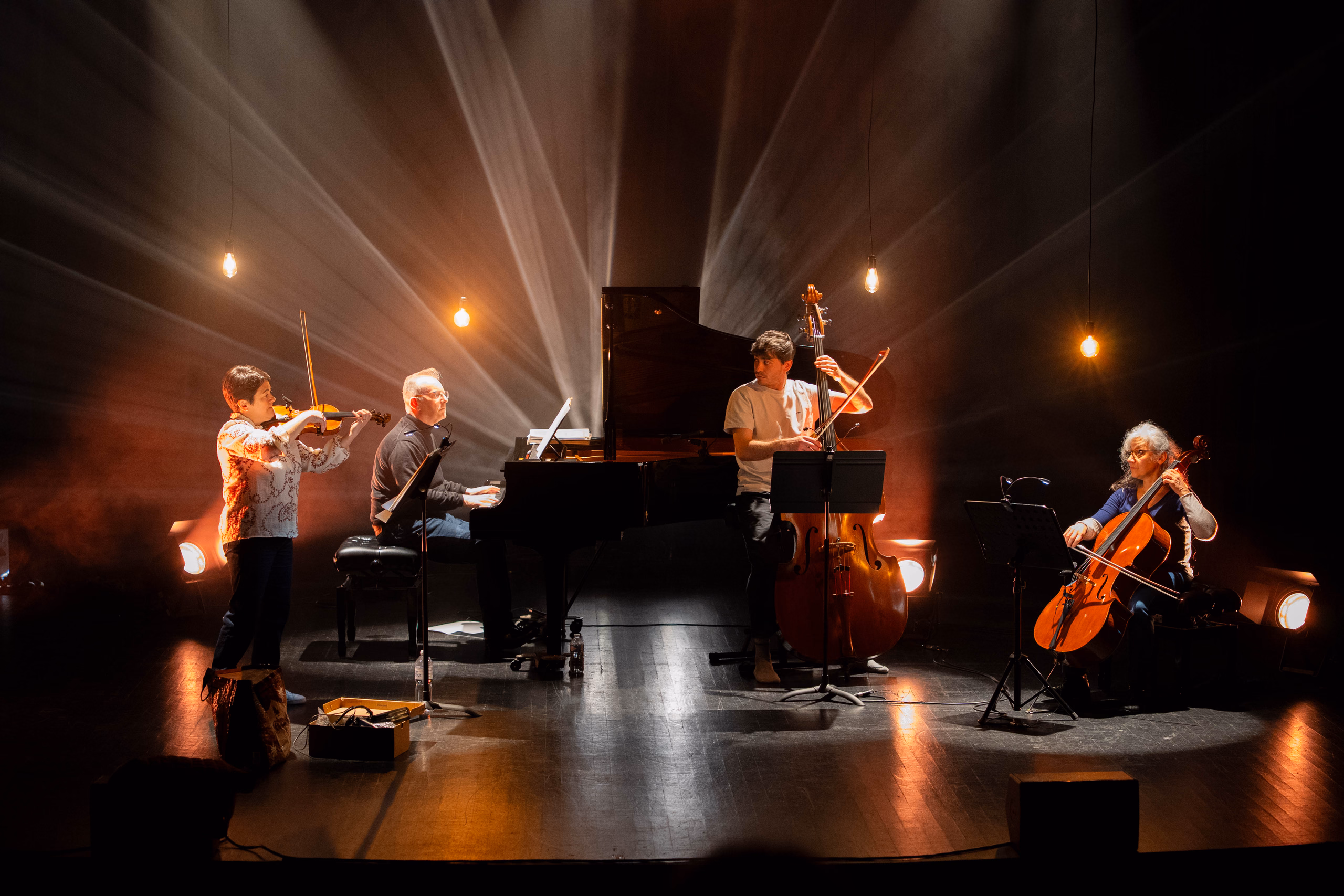 Quatuor sur scène (violon, piano à queue, contrebasse, violoncelle) sous des faisceaux de lumière chaude et des ampoules suspendues