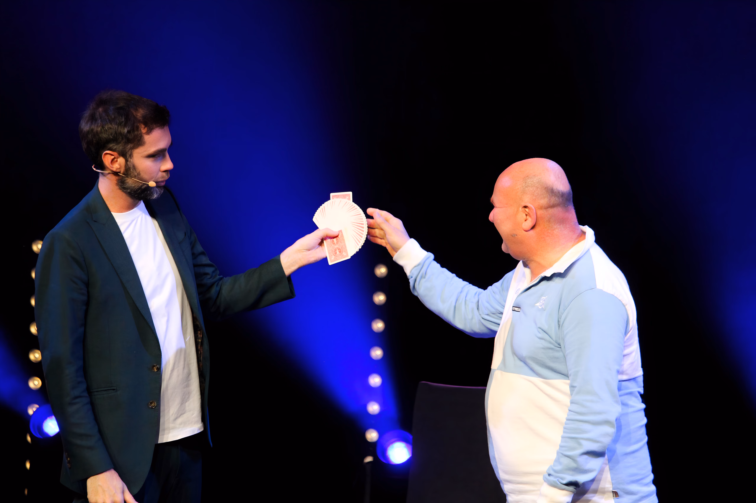 Tour de cartes sur scène : l'artiste présente un éventail de cartes à une personne du public, sous une lumière bleue